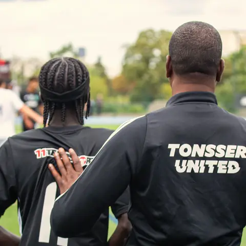 A Tonsser United coach with a hand on a player's back, watching the game together.